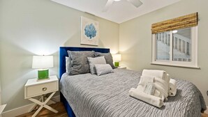 A bedroom with a bed, a ceiling fan, a window with blinds, a bedside table with a lamp, and a wall-mounted artwork. - Oceanfront Sunrises on Mitchelville Beach (Hilton Head Island)
