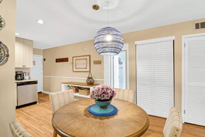A dining area with a round wooden table, chairs, a vase with flowers, a hanging light fixture, and a kitchen in the background. - Soundfront Stay Beach, Pools & More at The Spa (Hilton Head Island)