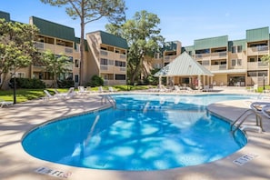 A swimming pool with lounge chairs, a covered seating area, and a building with multiple balconies.