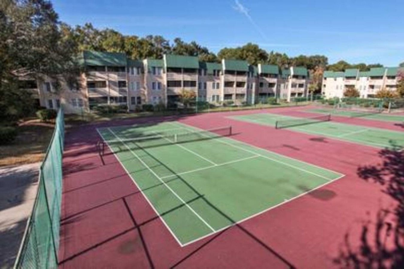 Una cancha de tenis con red, rodeada por una cerca, y contigua a un edificio con varios pisos y balcones.