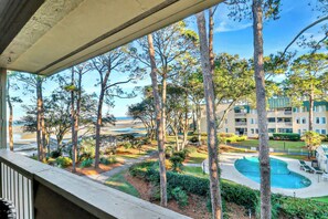 A pool area with a curved design, surrounded by trees and a building with balconies. - Soundfront Stay Beach, Pools & More at The Spa (Hilton Head Island)