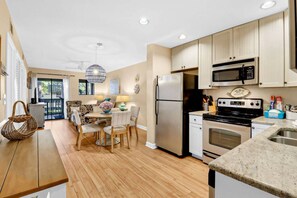 A modern kitchen with stainless steel appliances, a dining area with a round table and chairs, and a view through the glass door. - Soundfront Stay Beach, Pools & More at The Spa (Hilton Head Island)