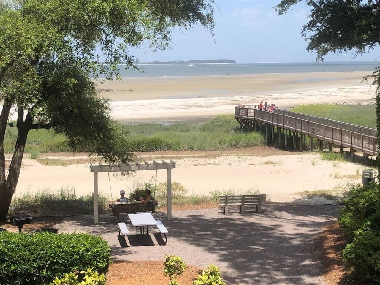 A beachside park with a wooden pier, picnic tables, and a covered seating area.