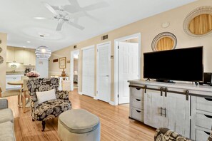 A living room with a sofa, ottoman, TV, and a dining area with a chandelier. - Soundfront Stay Beach, Pools & More at The Spa (Hilton Head Island)
