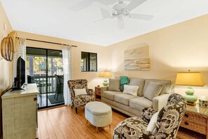 A living room with a sofa, armchairs, a coffee table, a TV, and a balcony with a view. - Soundfront Stay Beach, Pools & More at The Spa (Hilton Head Island)