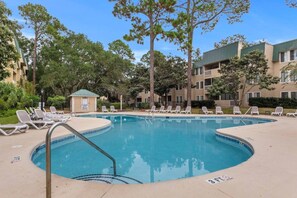 A swimming pool with lounge chairs and a small building, surrounded by trees and a multi-story building.