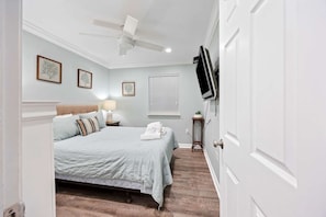Une chambre à coucher avec un lit, un ventilateur de plafond, un téléviseur fixé au mur et une fenêtre avec des stores.