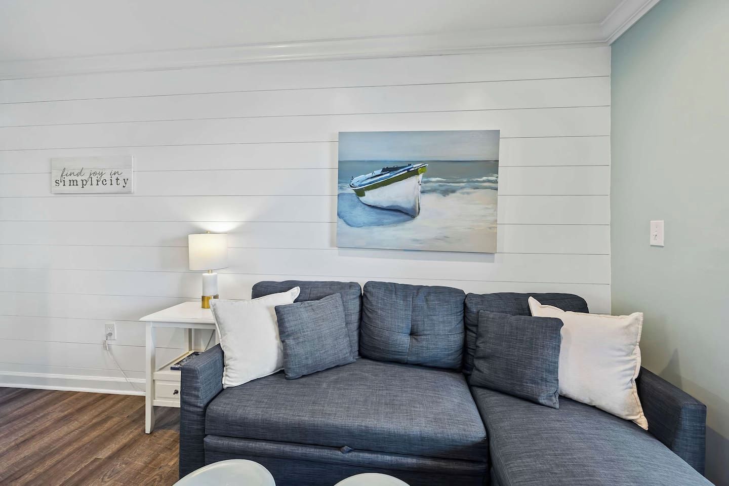 A modern living room with a grey sectional sofa, a small white side table with a lamp, and a painting of a boat on the wall.