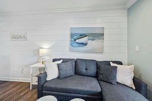 A modern living room with a grey sectional sofa, a small white side table with a lamp, and a painting of a boat on the wall.