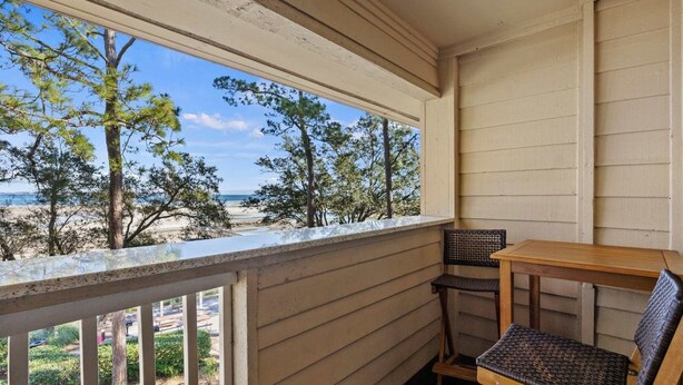 A balcony with a view of the beach and ocean, a wooden table, and two wicker chairs.