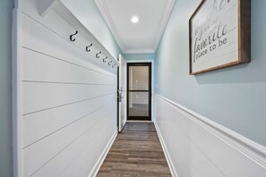 A hallway with a white wall, a wooden floor, and a door leading to a room. There is a framed quote on the wall.