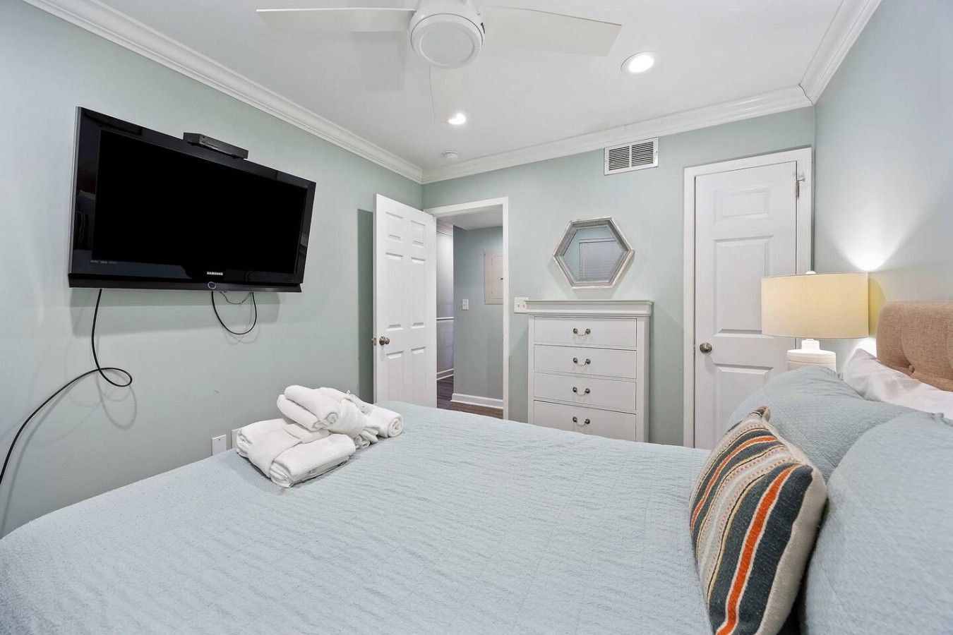 A bedroom with a bed, a television mounted on the wall, a dresser, a mirror, and a lamp.