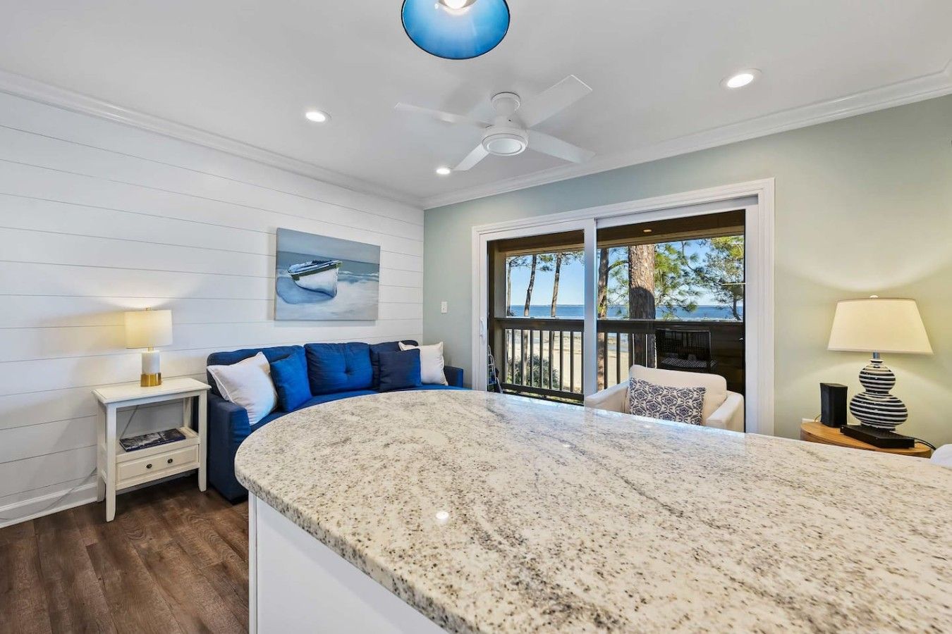 A modern kitchen with a granite countertop, a ceiling fan, and a view of the beach through large windows.