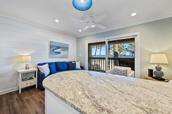 A modern kitchen with a granite countertop, a ceiling fan, and a view of the beach through large windows.