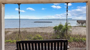Un banco de madera bajo una pérgola con vistas a una playa y un promontorio rocoso en el agua.