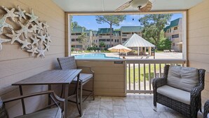 Un balcón con mesa y sillas, un sofá de mimbre, un ventilador de techo y vistas a una piscina y edificios de apartamentos.