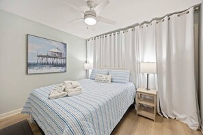 Une chambre Ă coucher avec un lit, un ventilateur de plafond, une table de chevet, une lampe, un tableau et des rideaux.