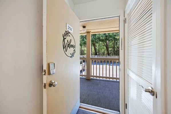A door with a 'Welcome' sign and a keypad lock, leading to a covered outdoor area with a view of trees and a parked car.