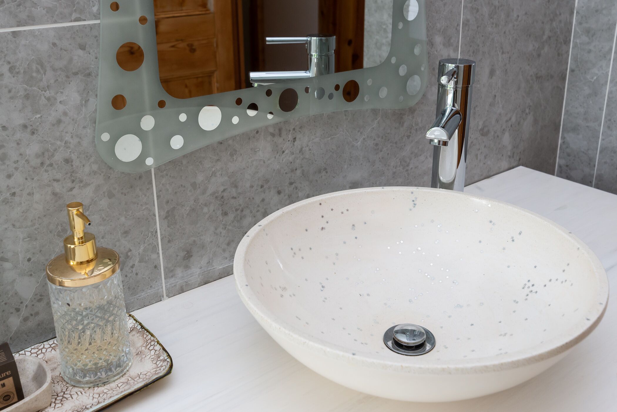A bathroom with a white sink, a gold soap dispenser, and a mirror with circular cutouts.