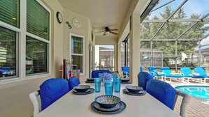Un patio techado con una mesa dispuesta para comer, sillas con cojines azules y una piscina al fondo.