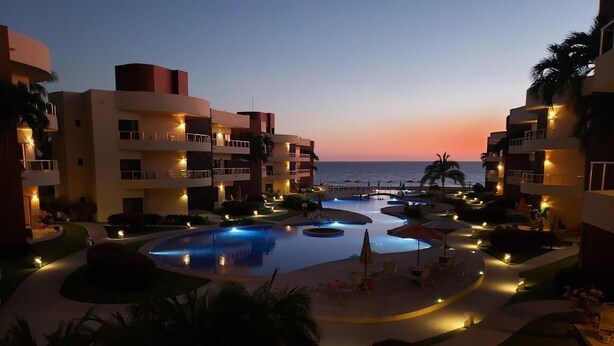 Zona de piscina en un resort con varias piscinas, palmeras y vistas a la playa al anochecer.