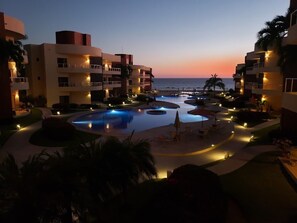 Un espace piscine d’un complexe hôtelier comprenant plusieurs piscines, des palmiers et une vue sur la plage au crépuscule.