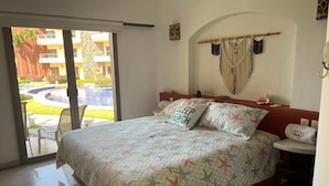 Un dormitorio con cama, una mesita de noche, ventana con vista a una piscina y edificios, y un colgador de macramé en la pared.
