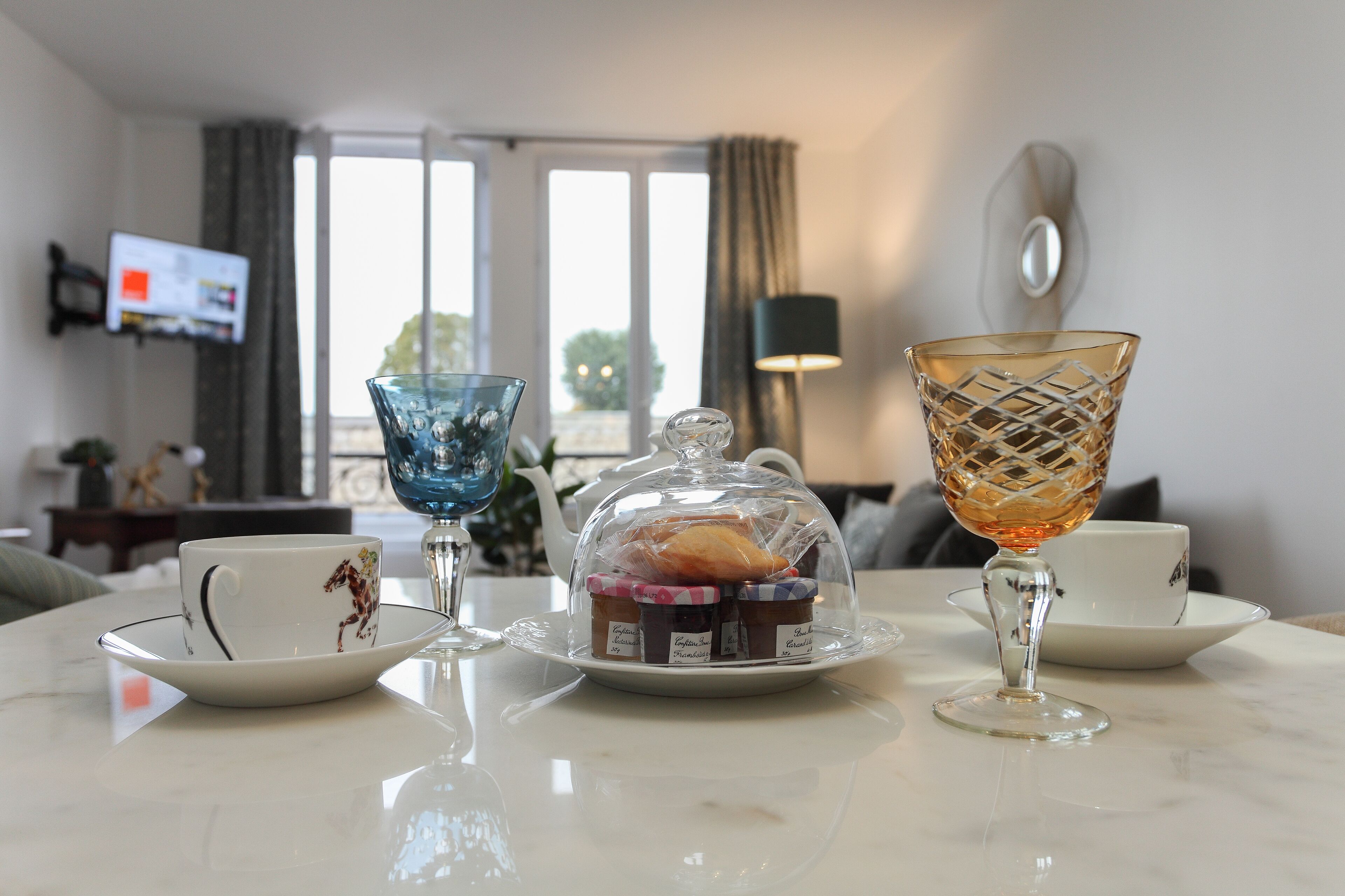 A table with a teapot, cups, and a glass on a marble surface in a room with a TV and curtains.