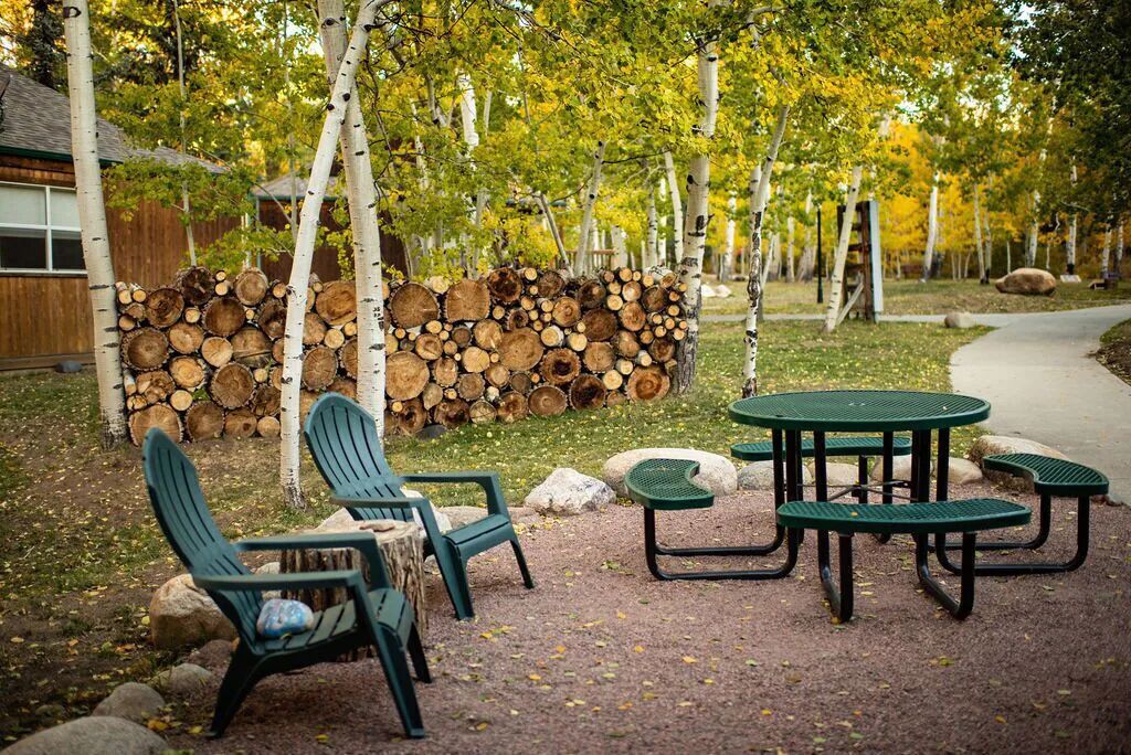 A picnic area with green metal chairs and tables, surrounded by a stack of firewood and birch trees.