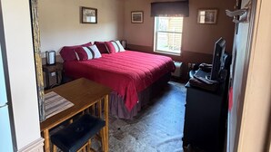 A room with a wooden ceiling, a bed with a red bedspread, a desk with a chair, a television, and a window with blinds. - Kara Creek Ranch - Double Bed Cabin with Kitchen #7 (Sundance)