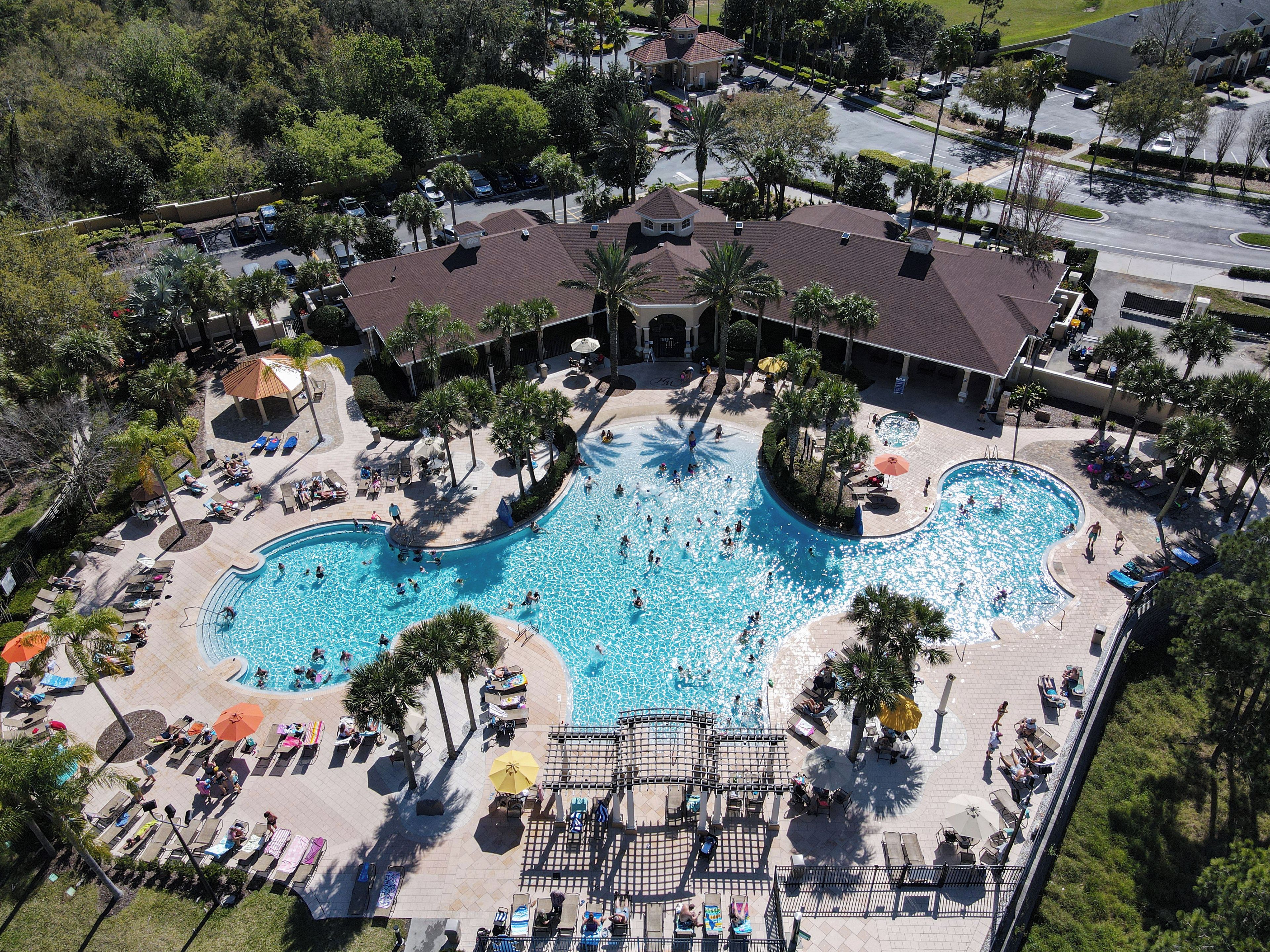 Eine Vogelperspektive zeigt ein Resort mit einem großen Schwimmbecken, das von Liegestühlen und Sonnenschirmen umgeben ist, sowie ein überdachtes Pavillon und einen Parkplatz.