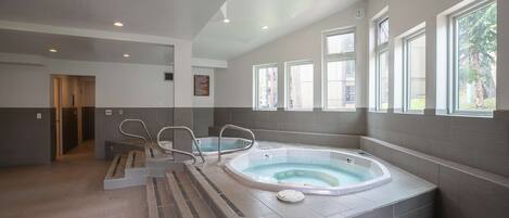 A modern spa room with a large hot tub, a shower area, and a view of the outdoors through large windows.