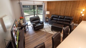 A modern living room with a leather sofa, a coffee table, and a TV. There is a window with curtains, a lamp, and a small table with flowers. - Gold Camp Miners' Inn Condo: Nice Location, Updates (Breckenridge)