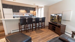 A modern kitchen with dark cabinets, a white countertop, and a bar area with stools. There is a television on a wooden stand and a leather ottoman in the living area. - Gold Camp Miners' Inn Condo: Nice Location, Updates (Breckenridge)