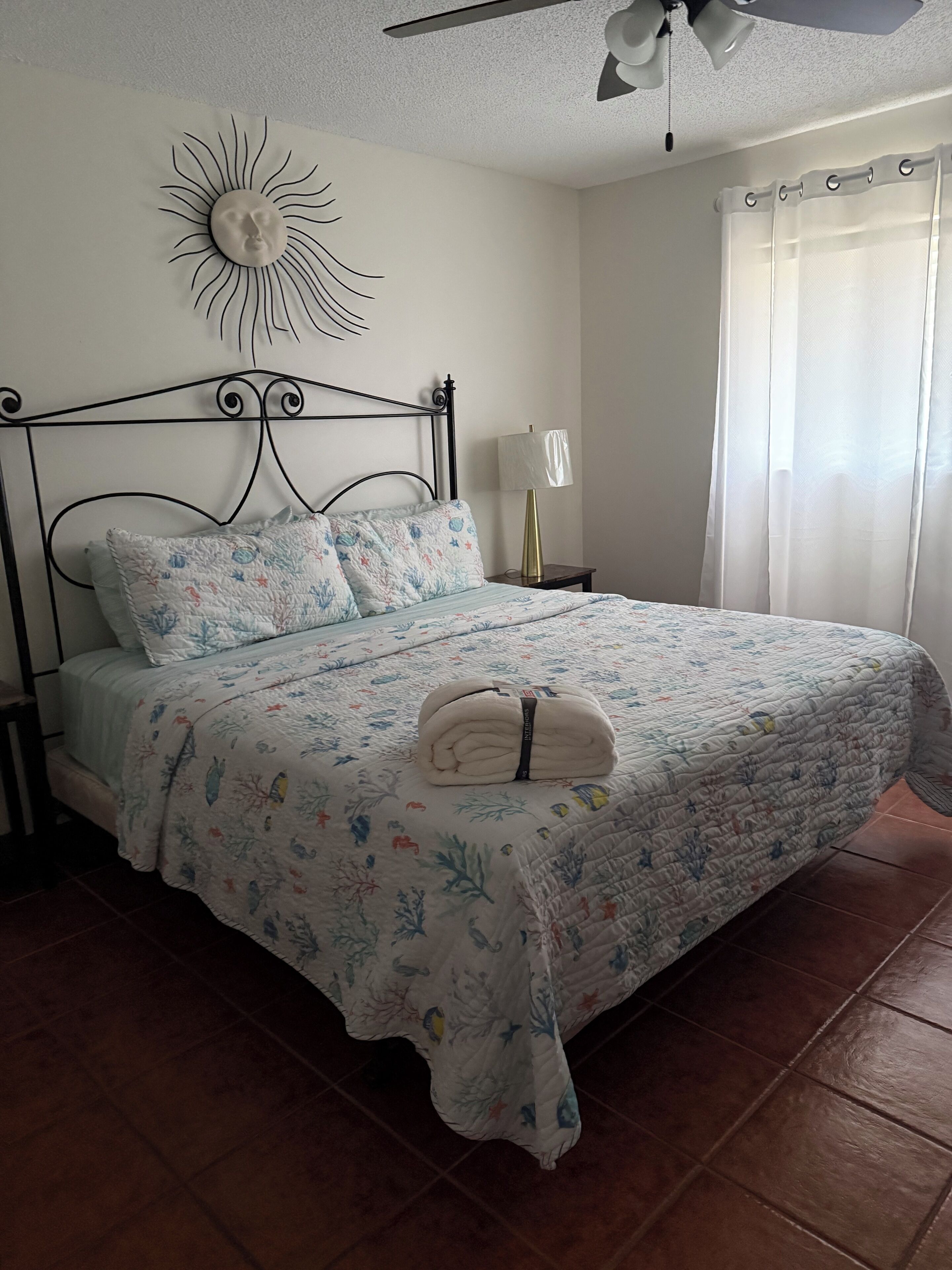 A neatly made bed with a floral-patterned bedspread, a decorative wall piece above the bed, and a ceiling fan.
