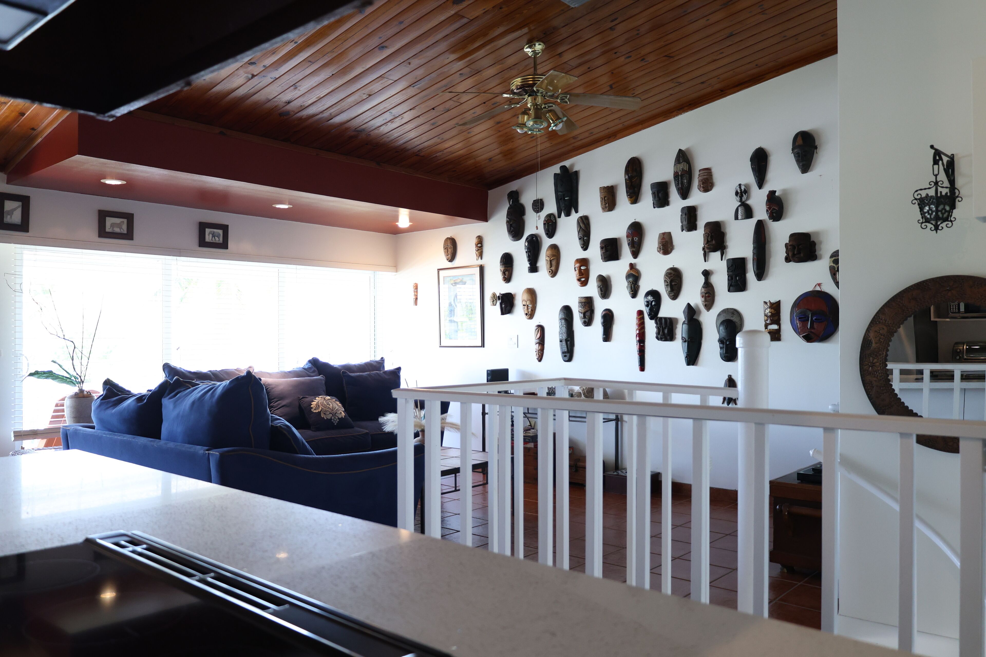 Una sala de estar con techo de madera, barandal blanco y una pared decorada con diversas máscaras.