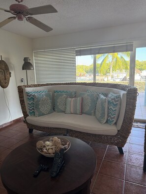 A room with a wicker sofa, a round table with a basket and remote controls, a ceiling fan, and a view of palm trees through the window.