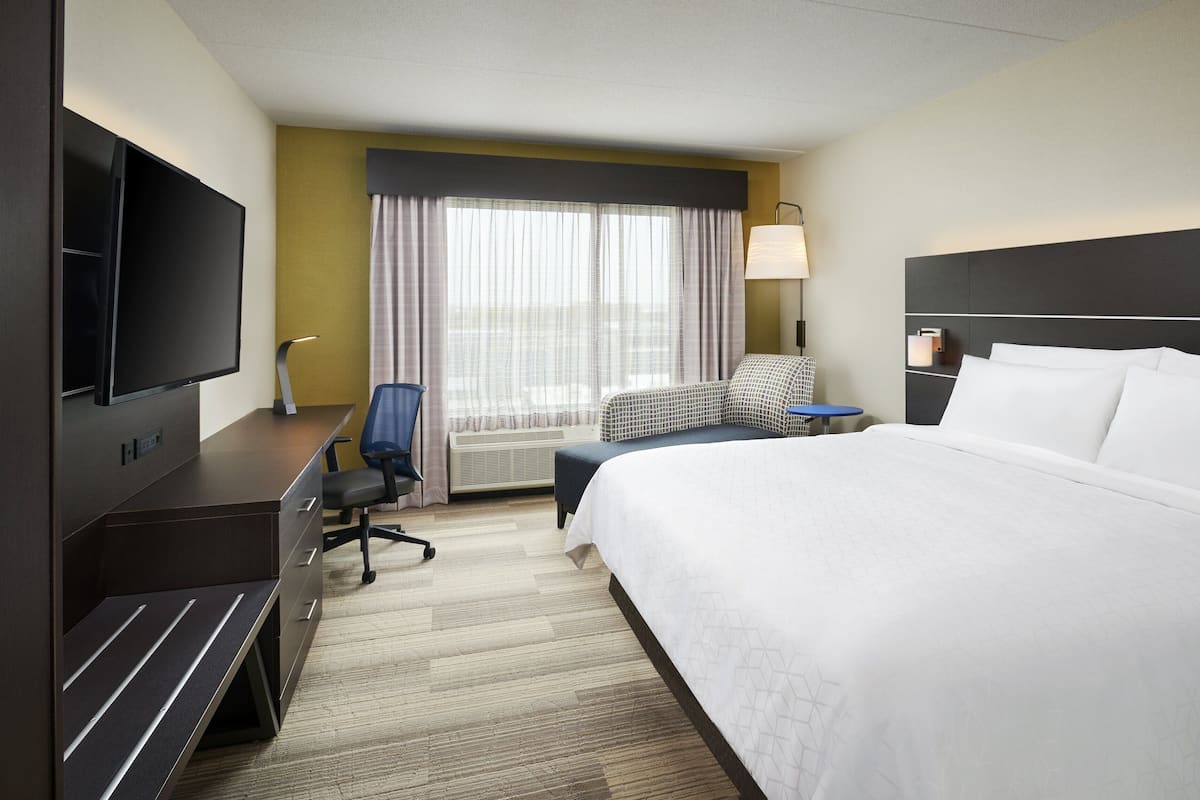 A hotel room with a large bed, a desk with a flat-screen TV, a chair, a small table, and a window with curtains.