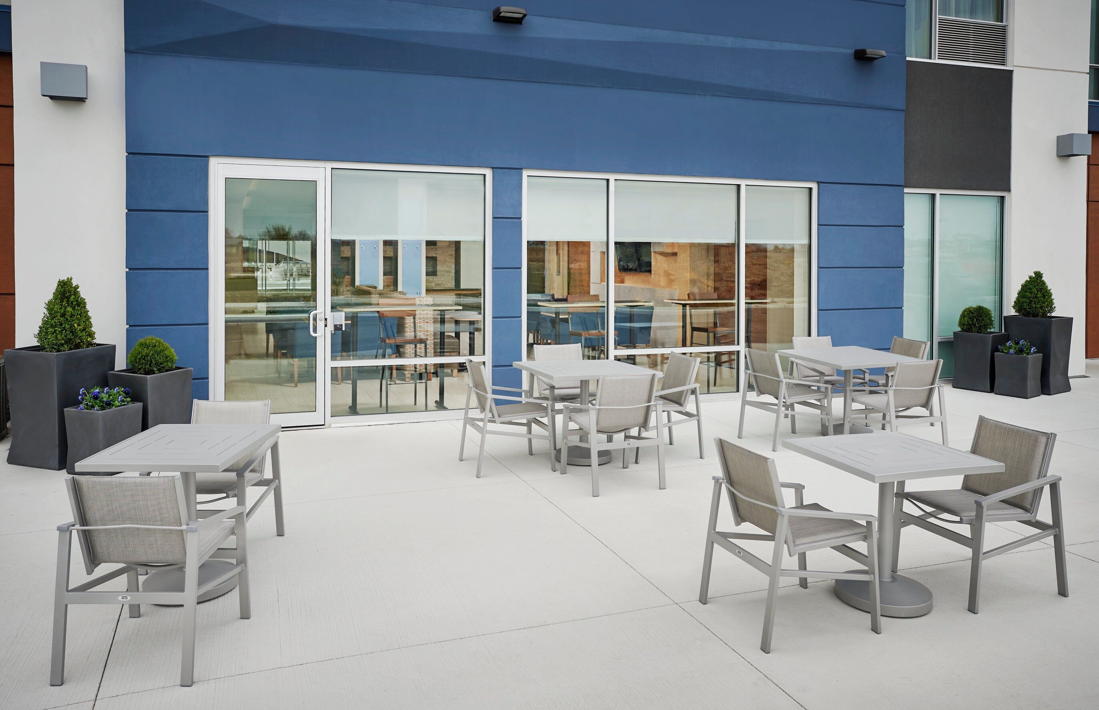 An outdoor patio area with tables and chairs, large windows, and potted plants.
