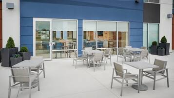 An outdoor patio area with tables and chairs, large windows, and potted plants.