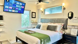 A modern bedroom with a bed, bedside tables, a TV mounted on the wall, a ceiling fan, and a window with a view of greenery.