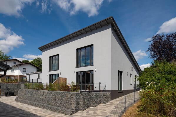 Ein modernes, zweistöckiges Haus mit einer Steinmauer im Vordergrund, großen Fenstern und wolkenlosem, blauem Himmel.