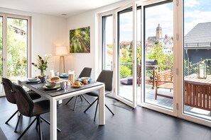 Ein moderner Essbereich mit einem für das Frühstück gedecktem Tisch, großen Fenstern mit Blick auf die Stadt und einem Balkon mit Sitzmöbeln.