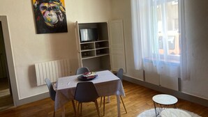 Ein Essbereich mit Tisch, Stühlen, einem kleinen runden Tisch, einem Heizkörper, einem Fernseher auf einem Regal und einem Fenster mit Vorhängen.
