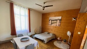 Ein Zimmer mit Holzboden, einem Bett mit weißer Bettdecke, einem Esstisch mit Stühlen, einem Deckenventilator und einem Bild an der Wand.