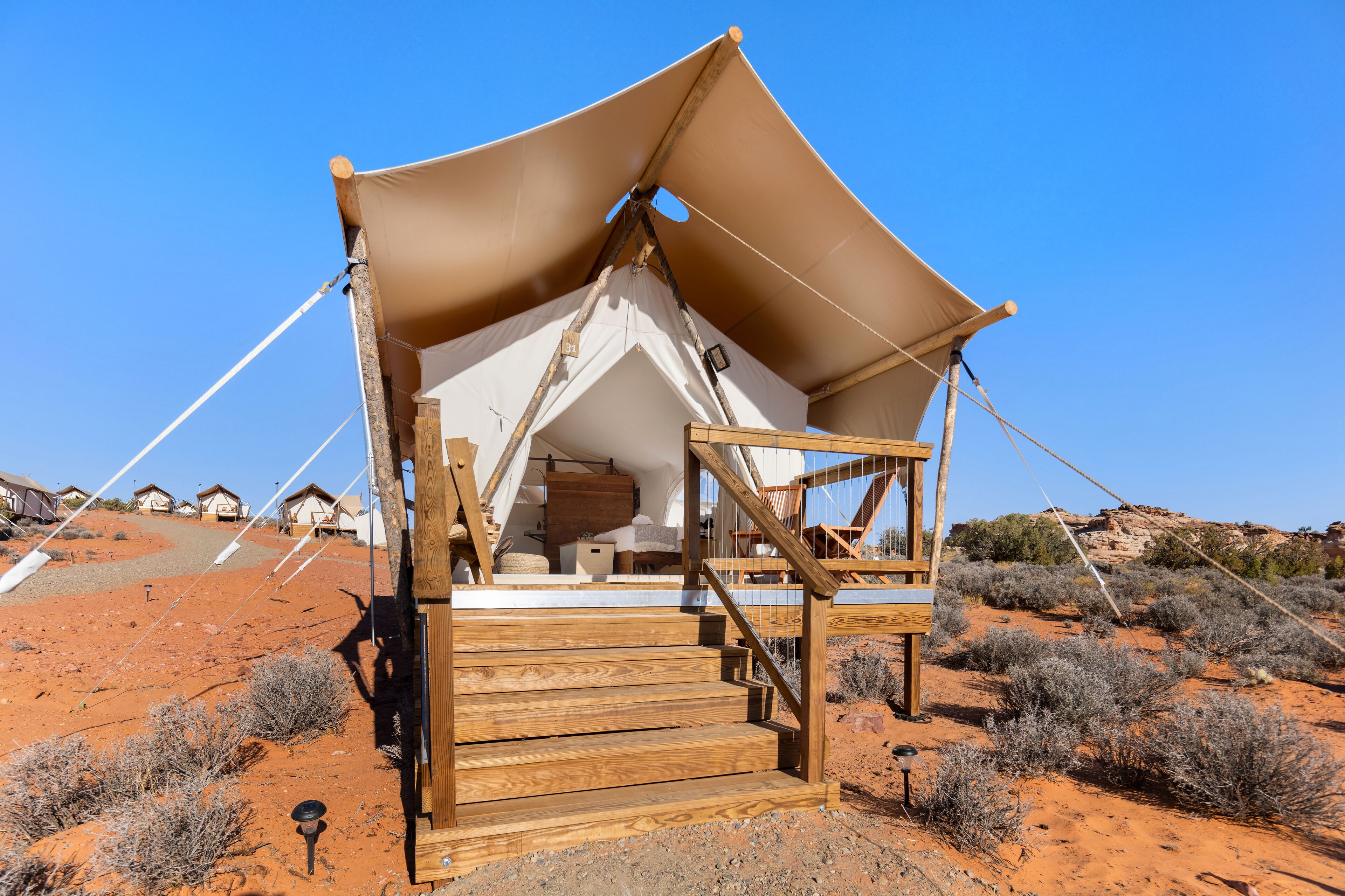 Una tienda de campaña en el desierto con escaleras de madera y un toldo grande, rodeada de vegetación escasa y casas a lo lejos.