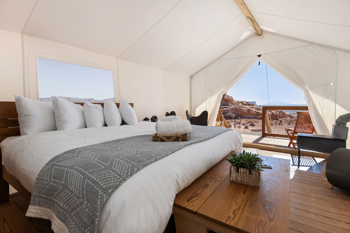 Een tentaccommodatie met een groot bed, houten vloer, een klein tafeltje met een plant en uitzicht op rotsachtig terrein.