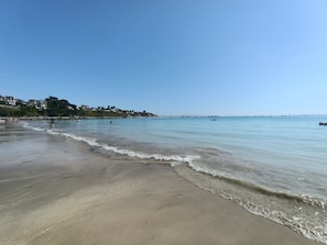 Une plage à l’eau claire, avec un rivage sablonneux et des bâtiments en arrière-plan.