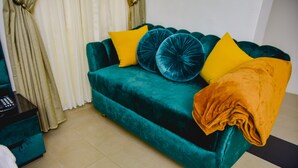 Un sofá color turquesa con cojines amarillos y azules, una manta dorada y una mesita auxiliar negra con un control remoto.