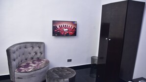 Una habitación moderna con un sillón capitoné, un otoman redondo, un televisor de pantalla plana en la pared y un armario oscuro.